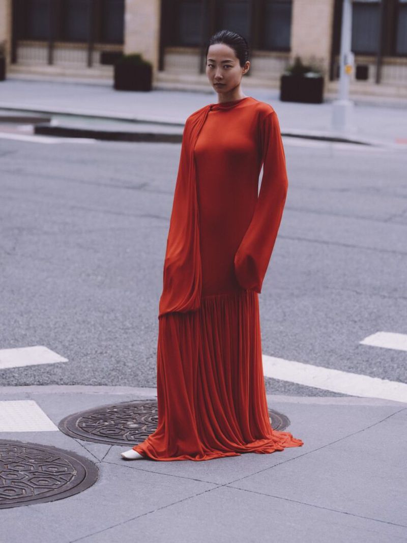 Asian woman in a long orange dress stands on a city street corner with muted buildings and asphalt ground under overcast daylight.