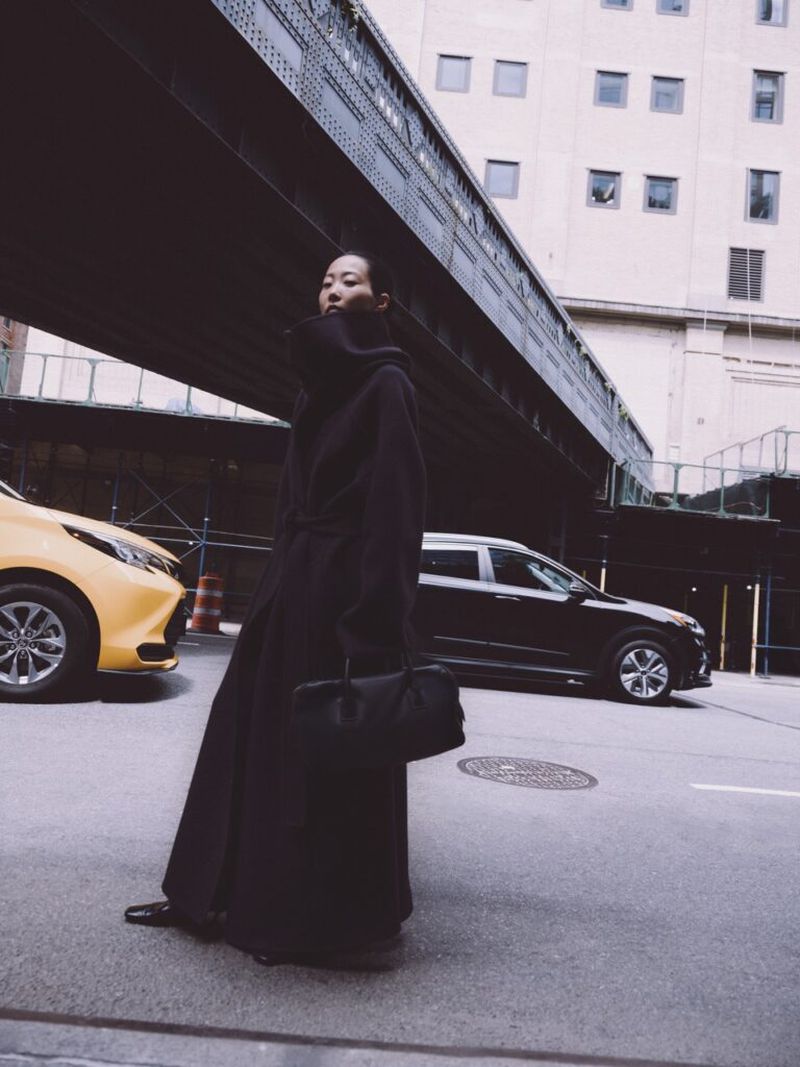 Woman in a long coat walks on a city street with buildings and an overpass, with yellow and black cars in the background.