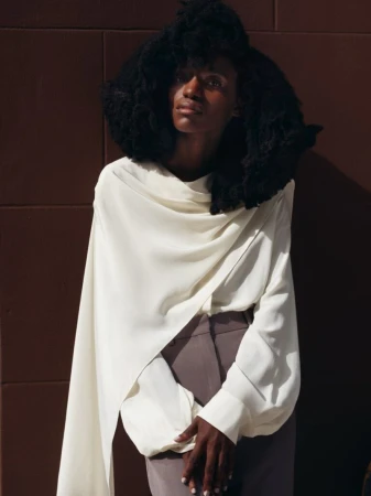 Black woman with voluminous natural hair wearing an asymmetrical draped off-white blouse and tailored trousers against a brown panel wall.