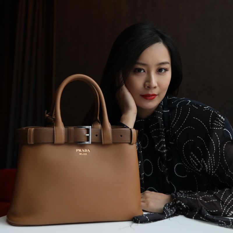 Elegant tan leather Prada handbag with belted detail, presented alongside a poised Asian woman in a patterned top and red lipstick.