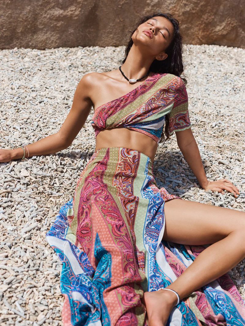 Woman in patterned one-shoulder crop top and long skirt reclines on pebble beach in warm sunlight. Fashion, summer, beach photography.