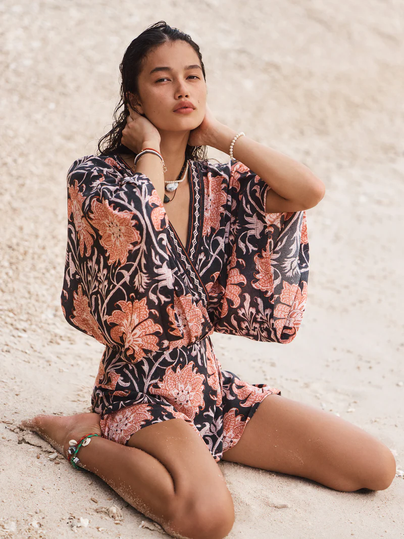 A tanned woman poses on a sandy beach, wearing a floral romper. Her hands rest on her neck, and she gazes off-camera with a neutral expression.