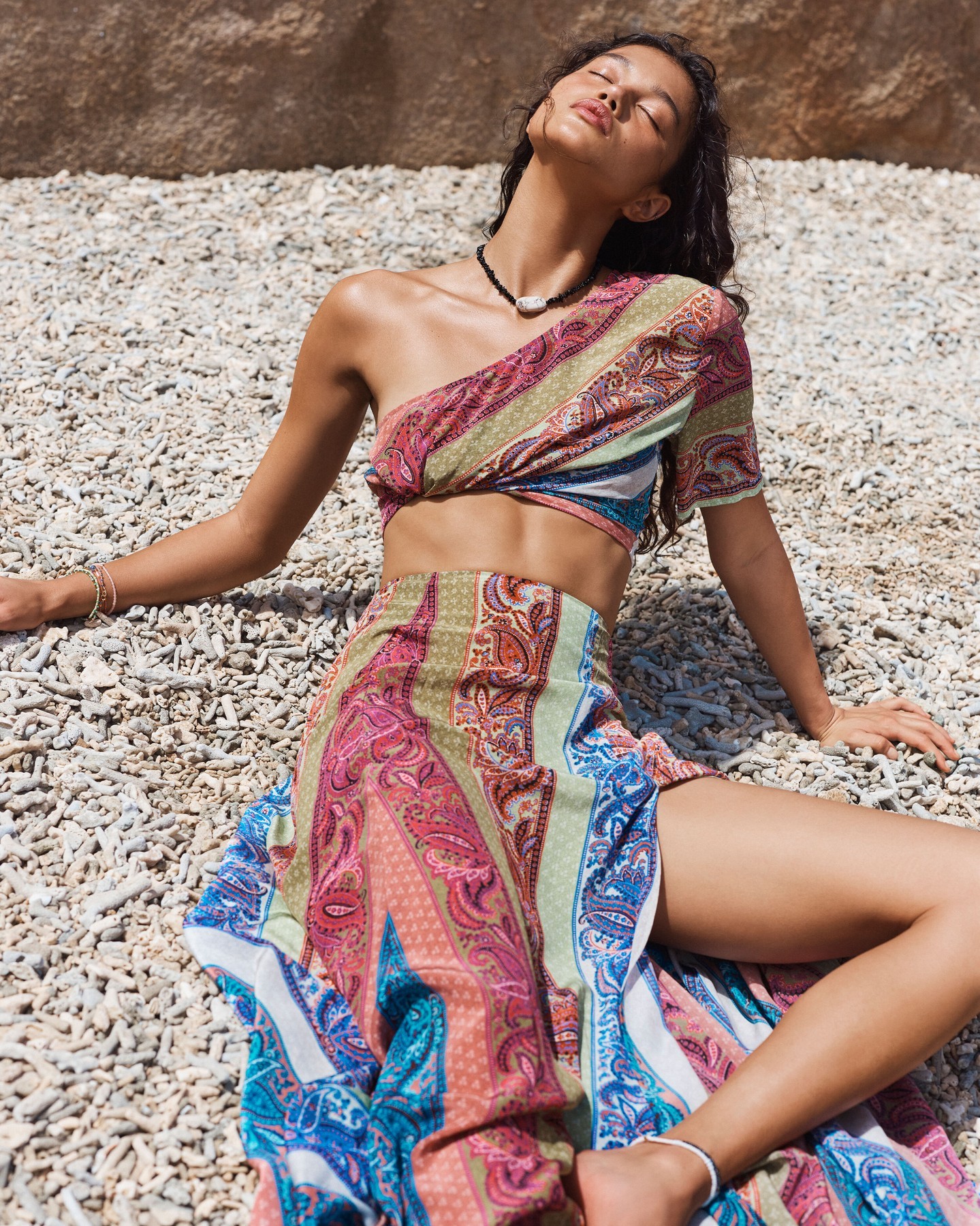 A woman in a paisley-patterned two-piece outfit lies on a stony beach with eyes closed enjoying the sun, showcasing a fashion and lifestyle aesthetic.