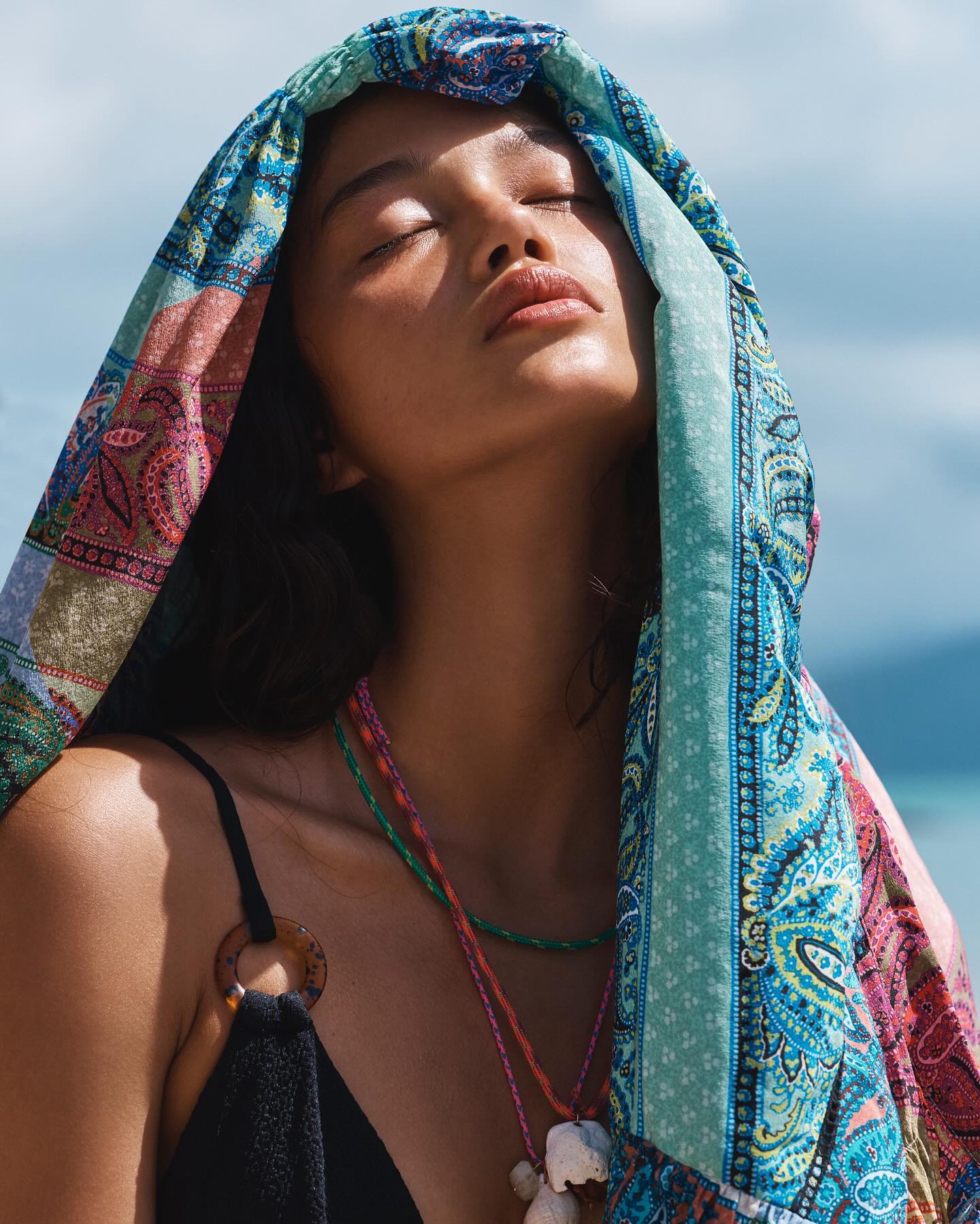Woman with closed eyes wearing a colorful headscarf and a black top with shell necklace on a sunny beach.