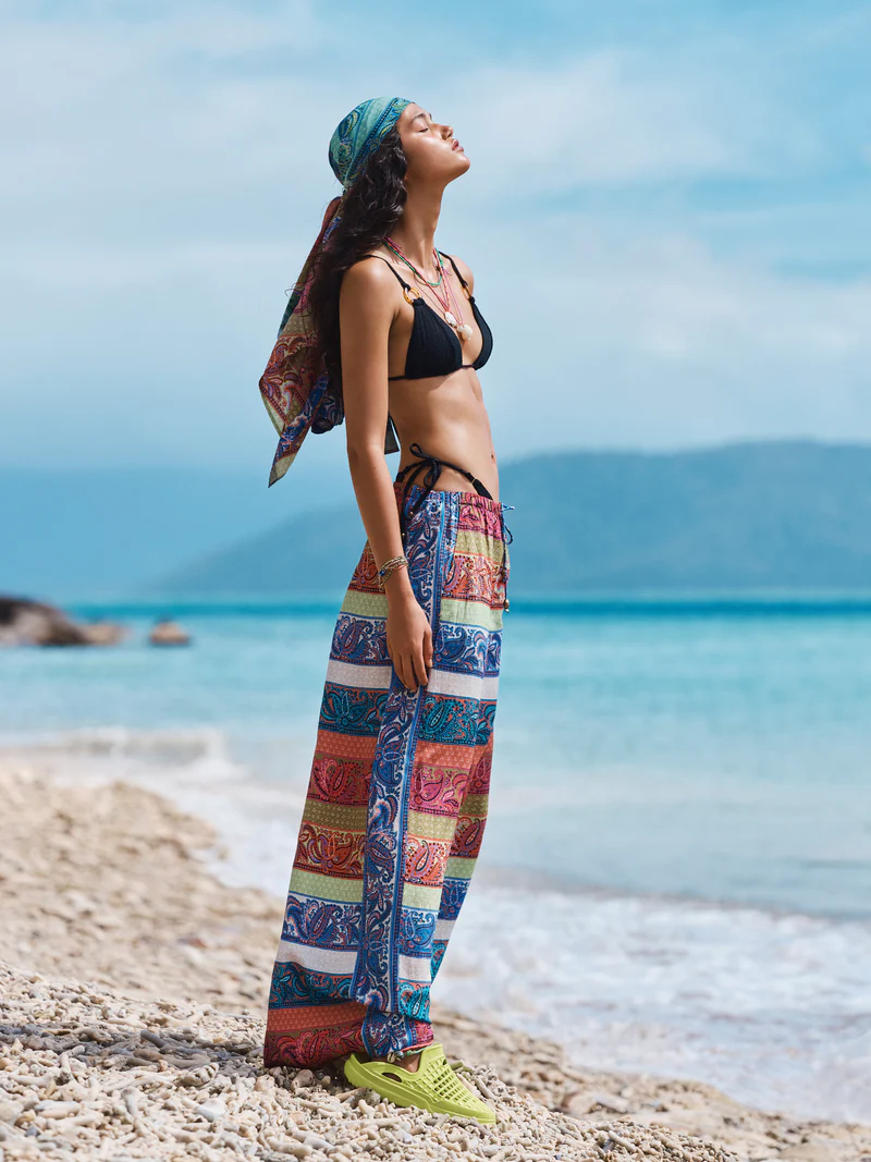 Woman in black bikini and patterned pants standing on a beach with turquoise water, wearing a headscarf, bathed in sunlight.