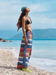 Model in patterned headscarf, black bikini top and colourful trousers standing on a sandy beach
