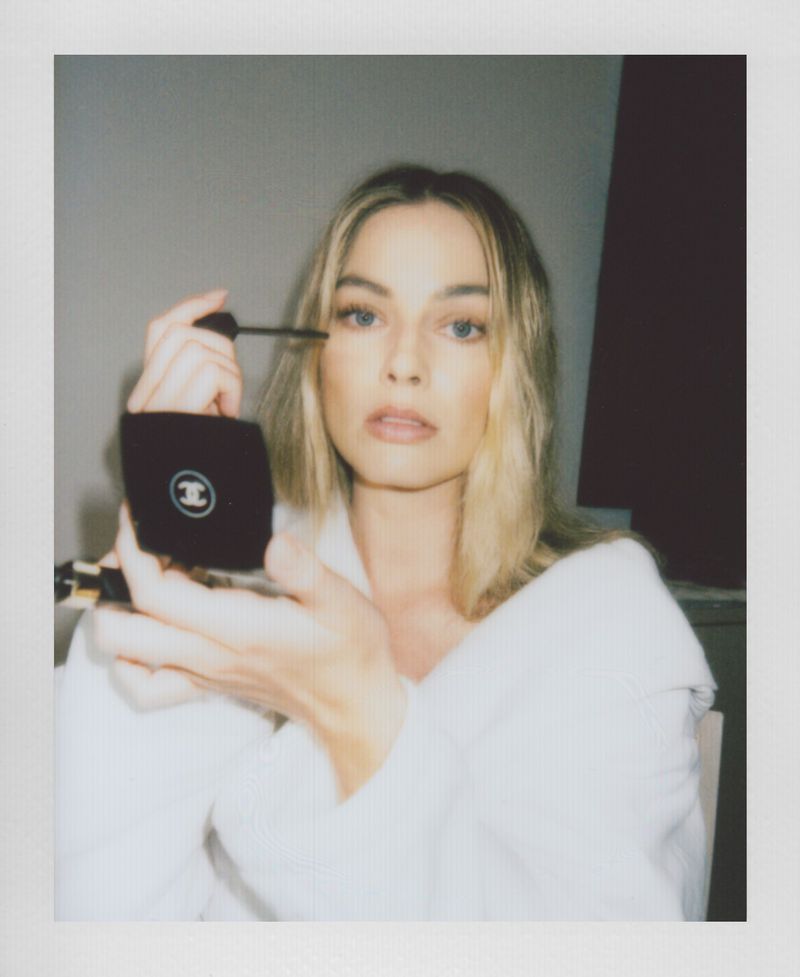 Close-up portrait of a blonde woman applying mascara, holding a Chanel compact mirror in an indoor setting with neutral background.