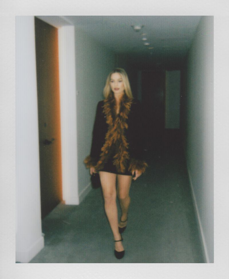 Stylish woman in a brown-feathered jacket and mini-dress walks down a hallway, highlighted by a warm-toned door, in a blurred photo.