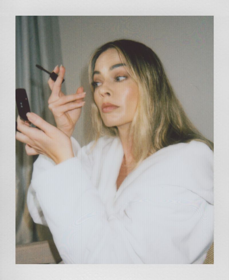 Blonde woman in a white robe applies mascara while holding a mirror, captured in a candid snapshot with soft lighting indoors.