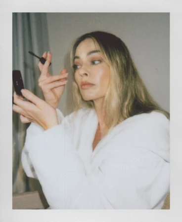 Blonde woman in a white robe applies mascara while holding a mirror, captured in a candid snapshot with soft lighting indoors.