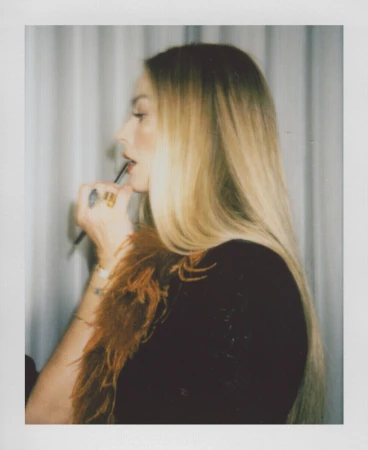 A woman with blonde hair, wearing a black dress with a feathered neckline, applies makeup with a brush in an indoor setting.