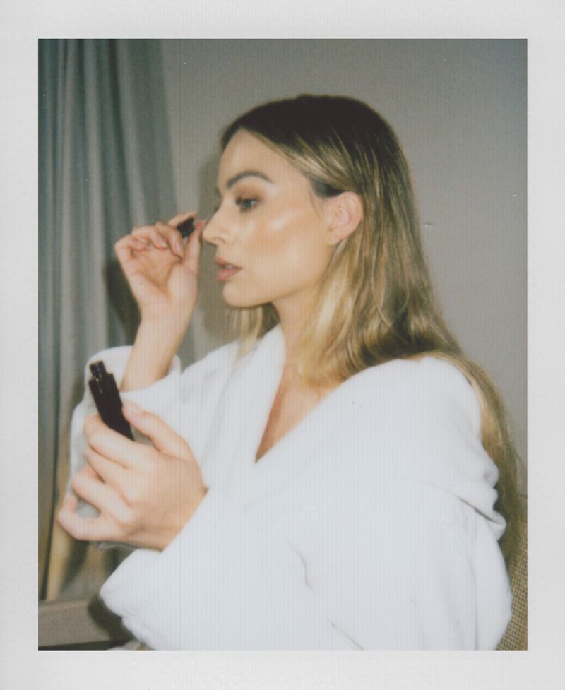 Portrait of adult female applying makeup with a brush indoors wearing a white robe against a neutral-colored wall in natural lighting.