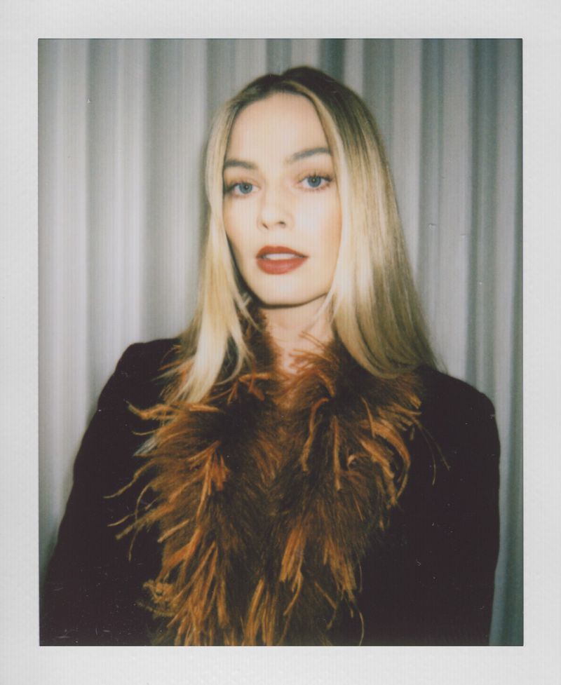Close-up shot of a blonde woman with dark red lipstick, wearing a dark jacket with orange feathered trim, in front of a white curtain.