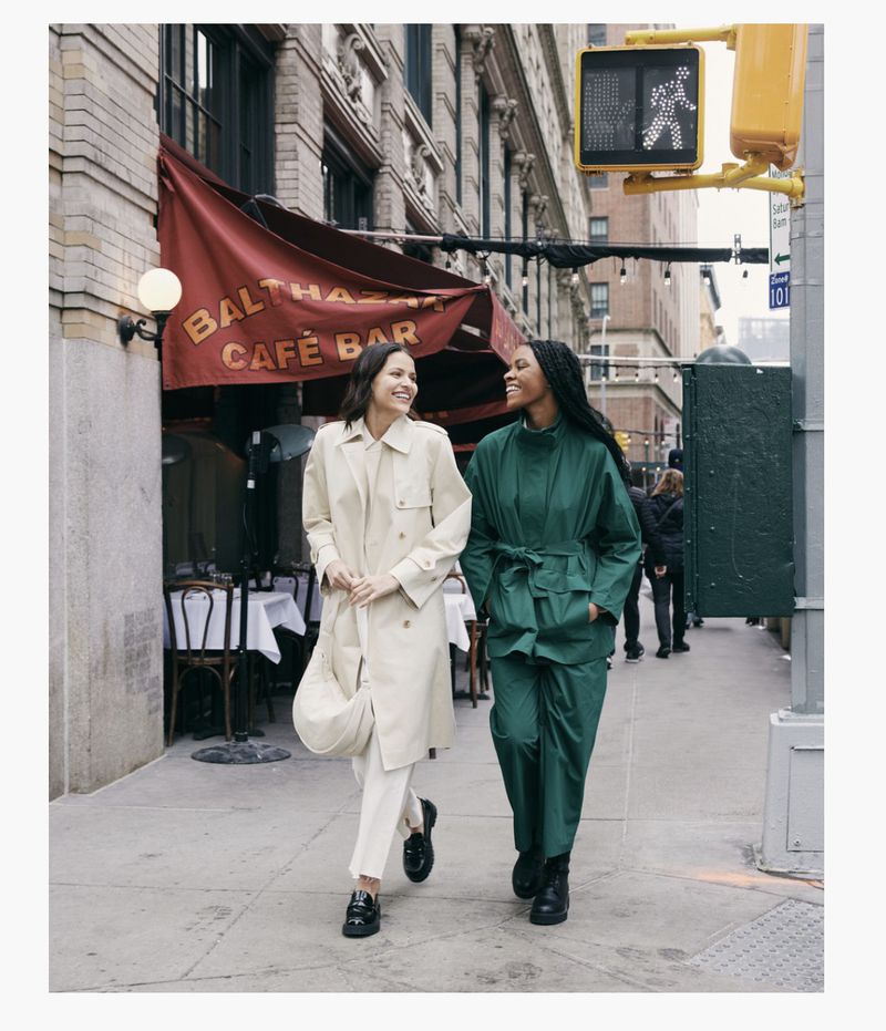 Two women walk side-by-side on a city sidewalk near an awning-covered restaurant, both smiling and wearing stylish, neutral-toned clothing.