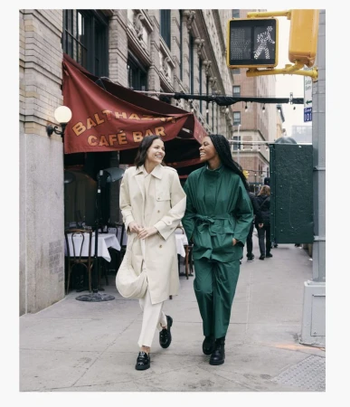 Two women walk side-by-side on a city sidewalk near an awning-covered restaurant, both smiling and wearing stylish, neutral-toned clothing.