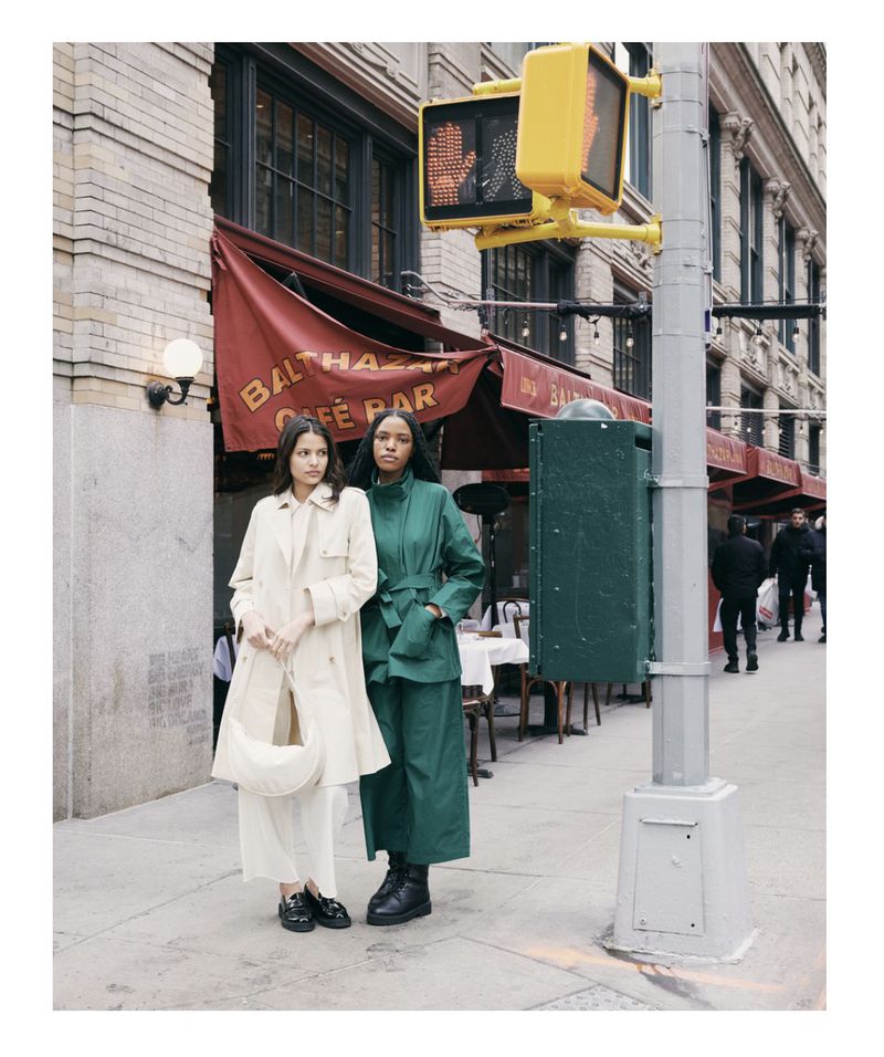 Two women in fashionable coats standing on a city street next to a traffic light and a restaurant awning, exuding urban style.