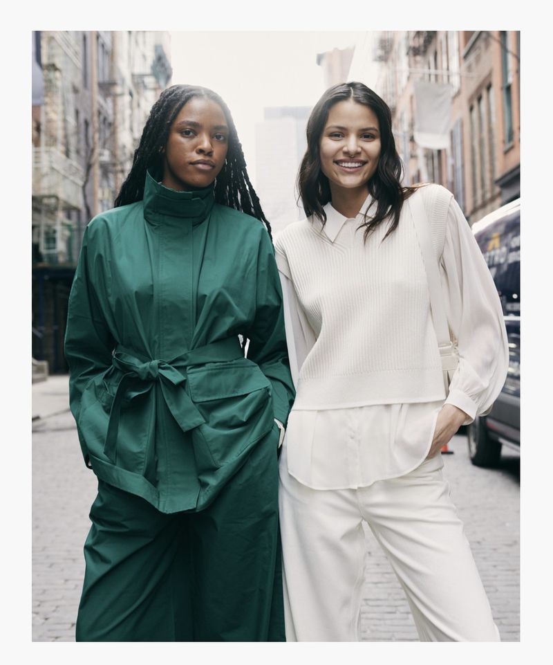Two women in stylish outerwear pose on a city street; one in a green coat, the other in a cream vest and pants ensemble.