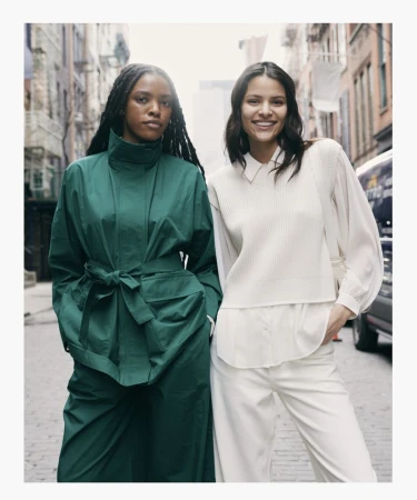 Two women in stylish outerwear pose on a city street; one in a green coat, the other in a cream vest and pants ensemble.