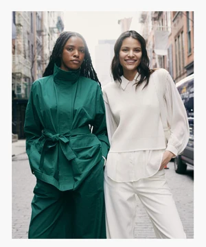 Two female models standing on an urban street, one in a green belted coat and the other in white knit vest and pants.