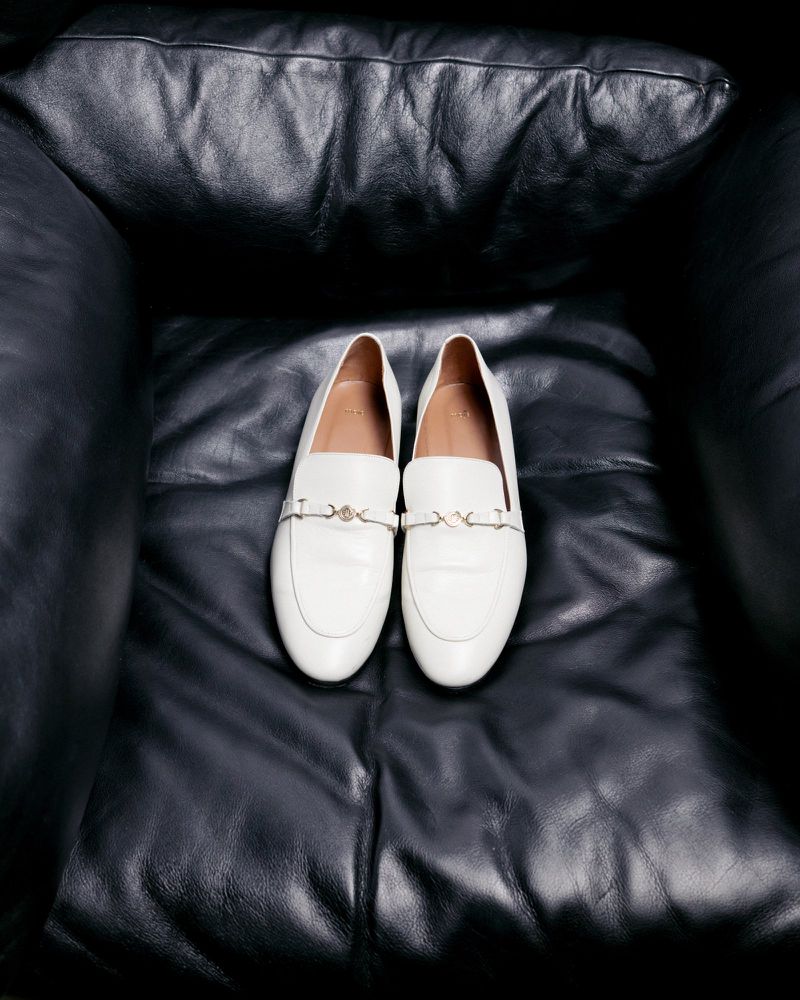 White leather loafers with gold hardware detail on a black leather chair. Fashion product photography highlighting the material and design.