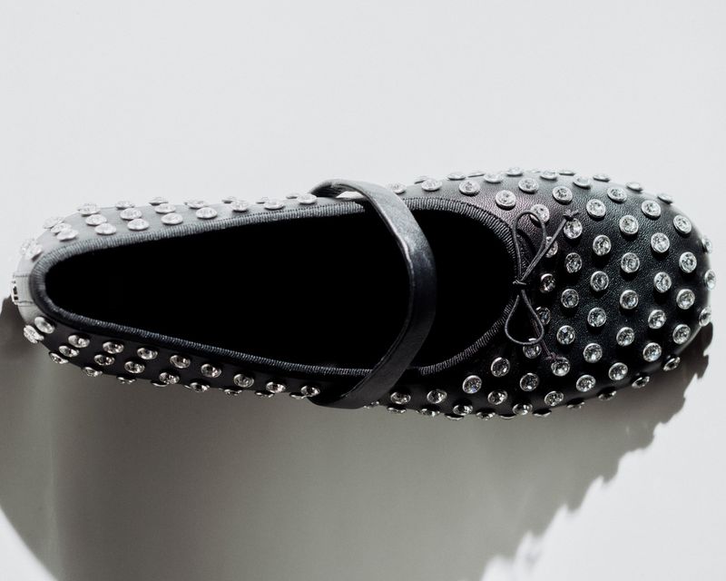 Black ballet flat shoe with strap and silver embellishments. Minimalist shoe photography against a bright white backdrop with soft lighting.