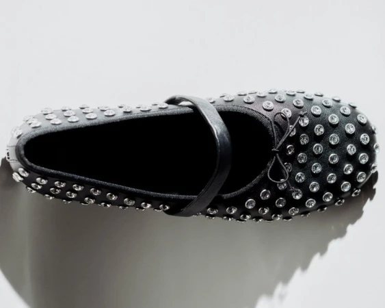 Black ballet flat shoe with strap and silver embellishments. Minimalist shoe photography against a bright white backdrop with soft lighting.