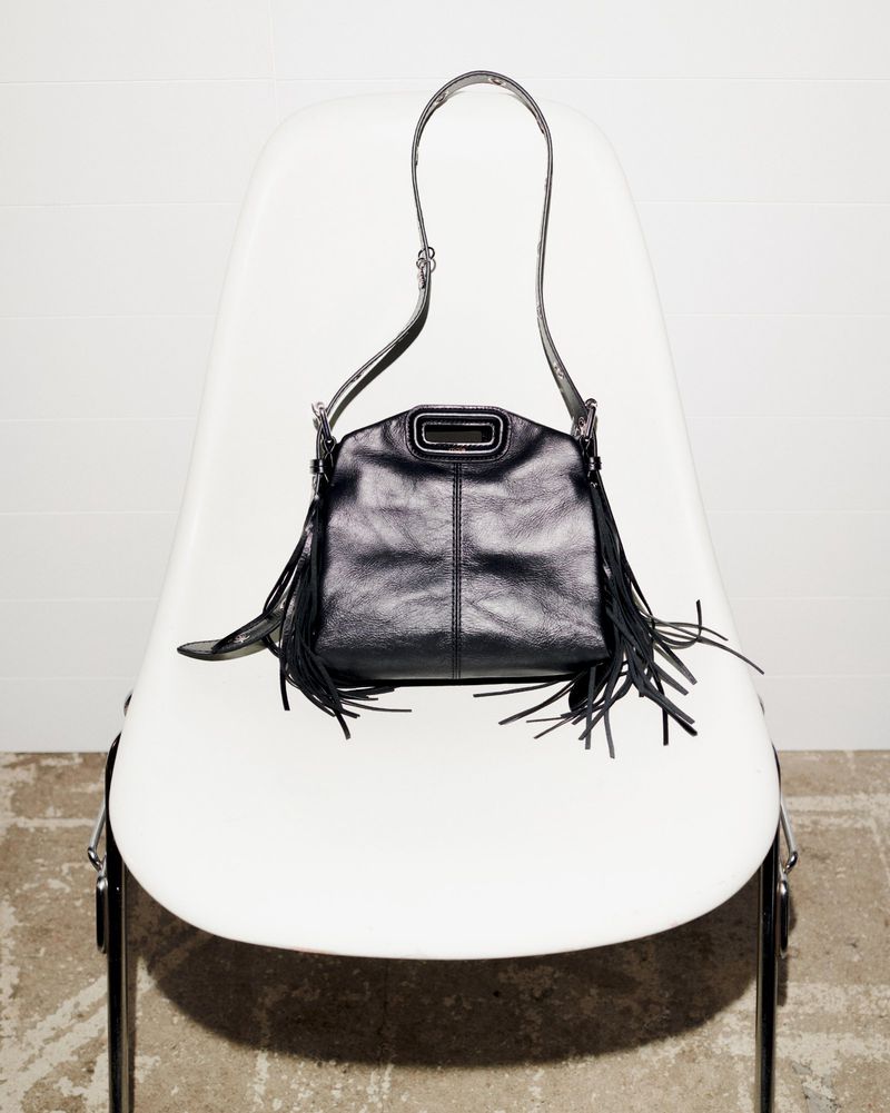 Black leather fringe purse with a top handle and shoulder strap resting on a white modern chair against a white wall and concrete floor.