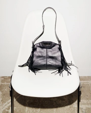 Black leather fringe purse with a top handle and shoulder strap resting on a white modern chair against a white wall and concrete floor.