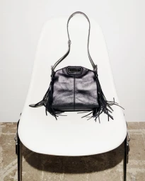 Black leather fringe shoulder bag resting on a white shell chair against a neutral wall