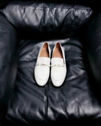 A pair of white leather loafers rests on a plush black leather armchair for Maje digital.