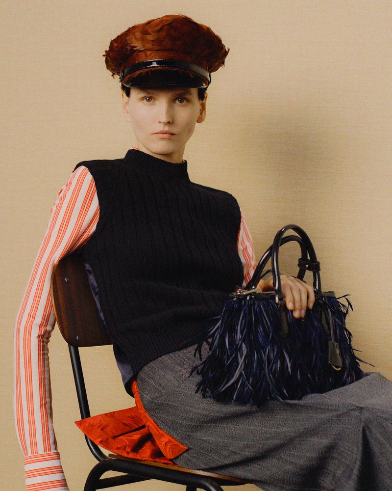 Indoor shot features a woman on a chair, wearing a striped shirt, vest, hat, and skirt, holding a handbag with feather details.