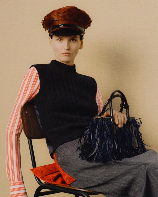 Indoor shot features a woman on a chair, wearing a striped shirt, vest, hat, and skirt, holding a handbag with feather details.