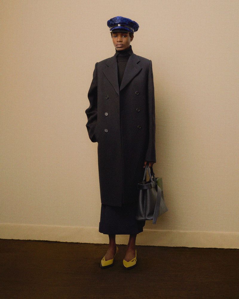 Elegant woman in a navy overcoat, matching skirt, blue cap, and yellow heels posing against a beige wall with a dark brown floor.