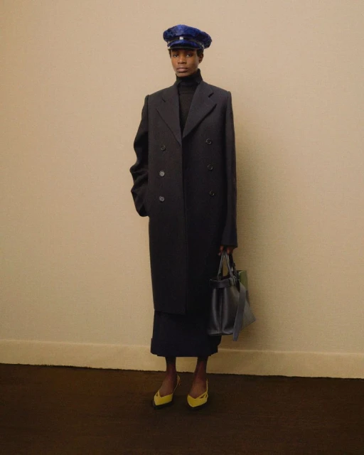 Elegant woman in a navy overcoat, matching skirt, blue cap, and yellow heels posing against a beige wall with a dark brown floor.