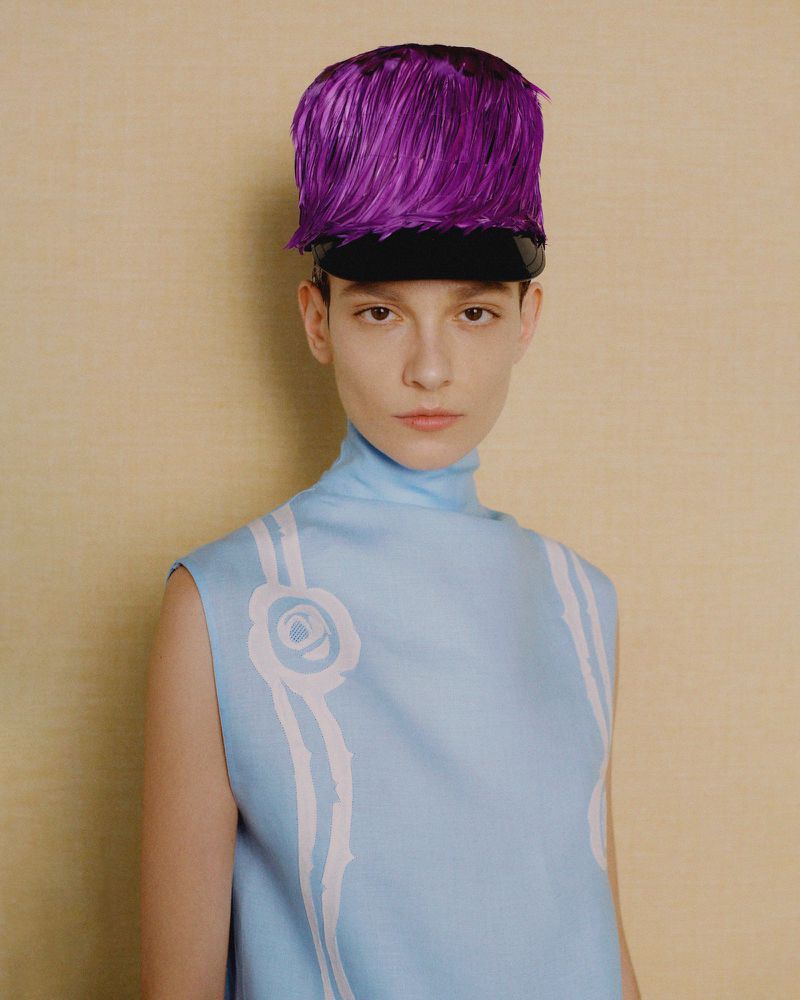 Portrait of a woman in a sleeveless light blue top and a purple feathered hat, posing against a neutral background in a studio setting.