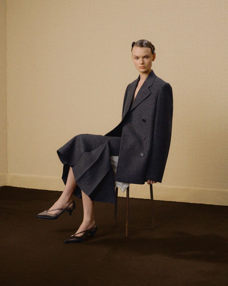 A woman sits on a stool indoors wearing a gray double-breasted suit and black kitten heels against a tan backdrop.