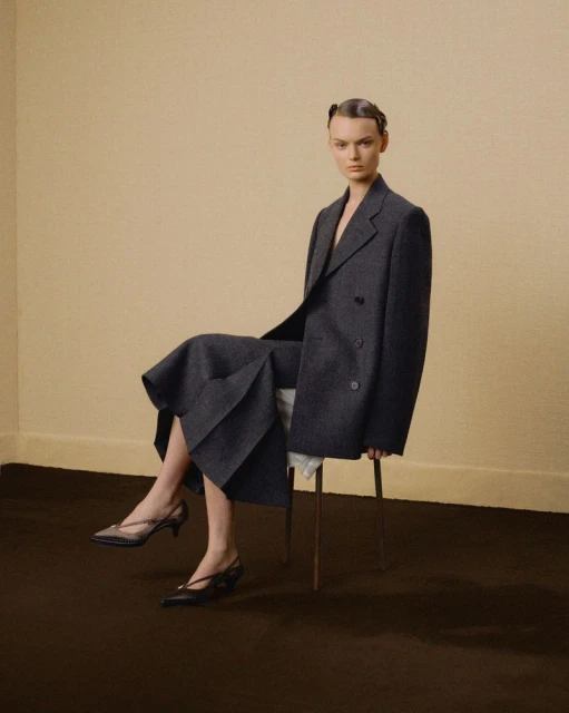 A woman sits on a stool indoors wearing a gray double-breasted suit and black kitten heels against a tan backdrop.