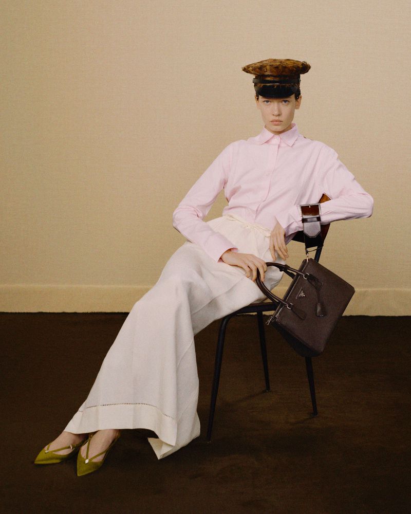 Fashion editorial image of a woman in a pink shirt, white trousers, and a cap, seated in a chair, holding a dark handbag indoors.