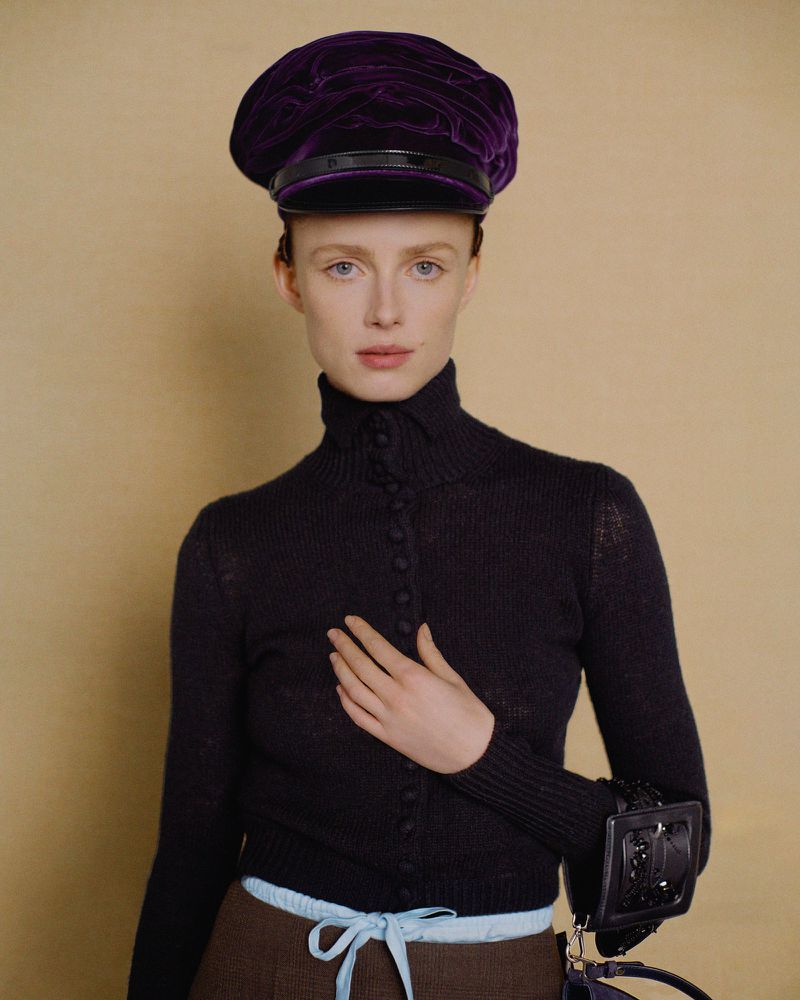 A young woman poses in a fashionable black turtleneck sweater and purple hat with a neutral background, showcasing a contemporary look.