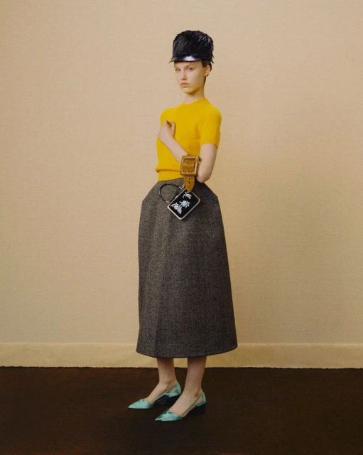 Woman poses in yellow top, grey skirt, cap, and heels against a neutral wall and brown floor, showcasing a vintage-inspired fashion style.