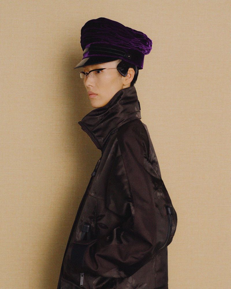 Stylish portrait of an Asian adult female wearing a dark jacket, glasses, and a purple hat, standing against a plain beige background.