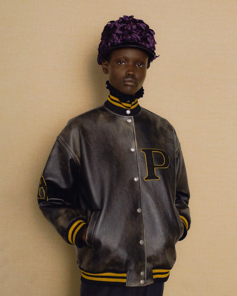 Portrait of a stylish young African woman wearing a black leather letterman jacket, a purple floral cap, and a black turtleneck.