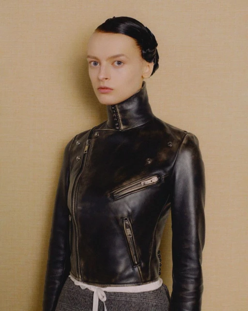 Portrait of a fair-skinned adult female with slicked-back hair wearing a black leather motorcycle jacket against a neutral background.
