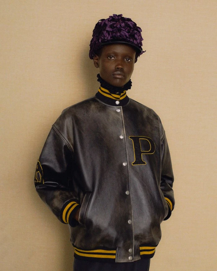 Model wearing a dark letterman jacket with yellow stripes and a textured purple floral cap for Prada Fall/Winter 2024.