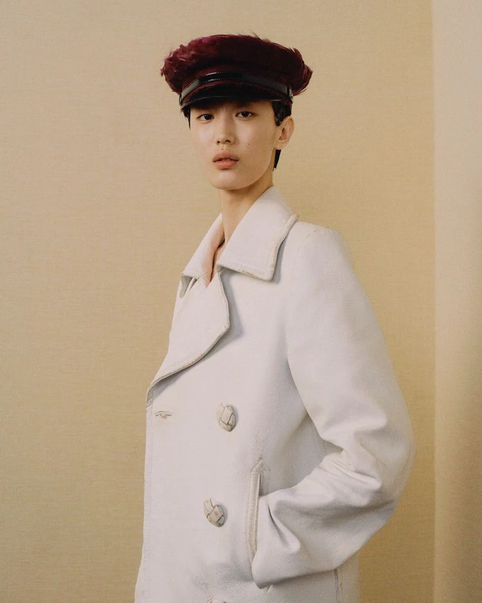 Model wearing a white double-breasted coat and dark maroon cap against a neutral beige wall for Prada.