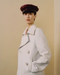 Model wearing a white double-breasted coat and dark maroon cap against a neutral beige wall for Prada.