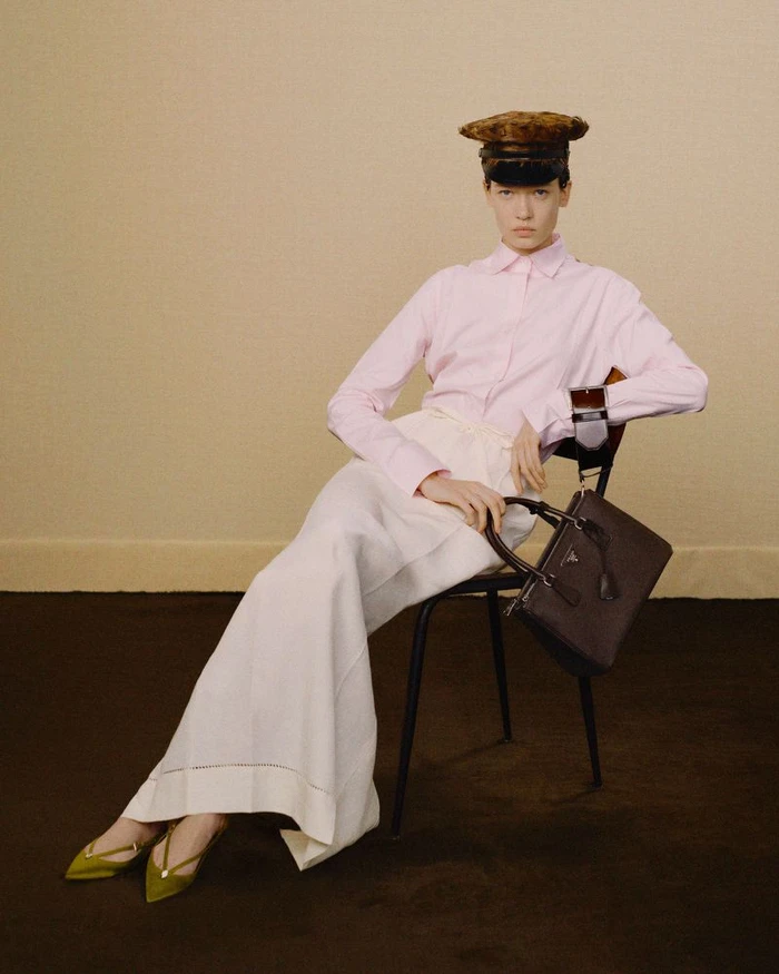 Model seated on chair wearing a pink button-up shirt, white skirt, patterned cap, and holding a dark handbag against a neutral background
