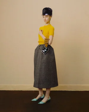 Model in yellow top and grey skirt with cap and handbag against neutral background