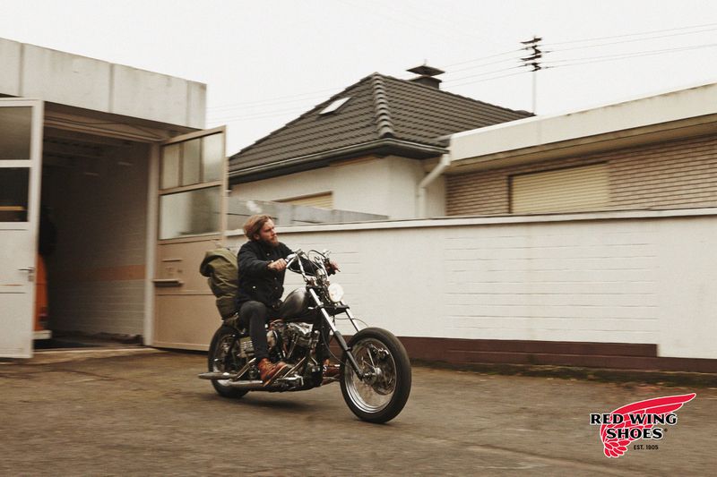 Adult male riding a vintage motorcycle on a concrete lot with a building and house in the background, promoting Red Wing Shoes.