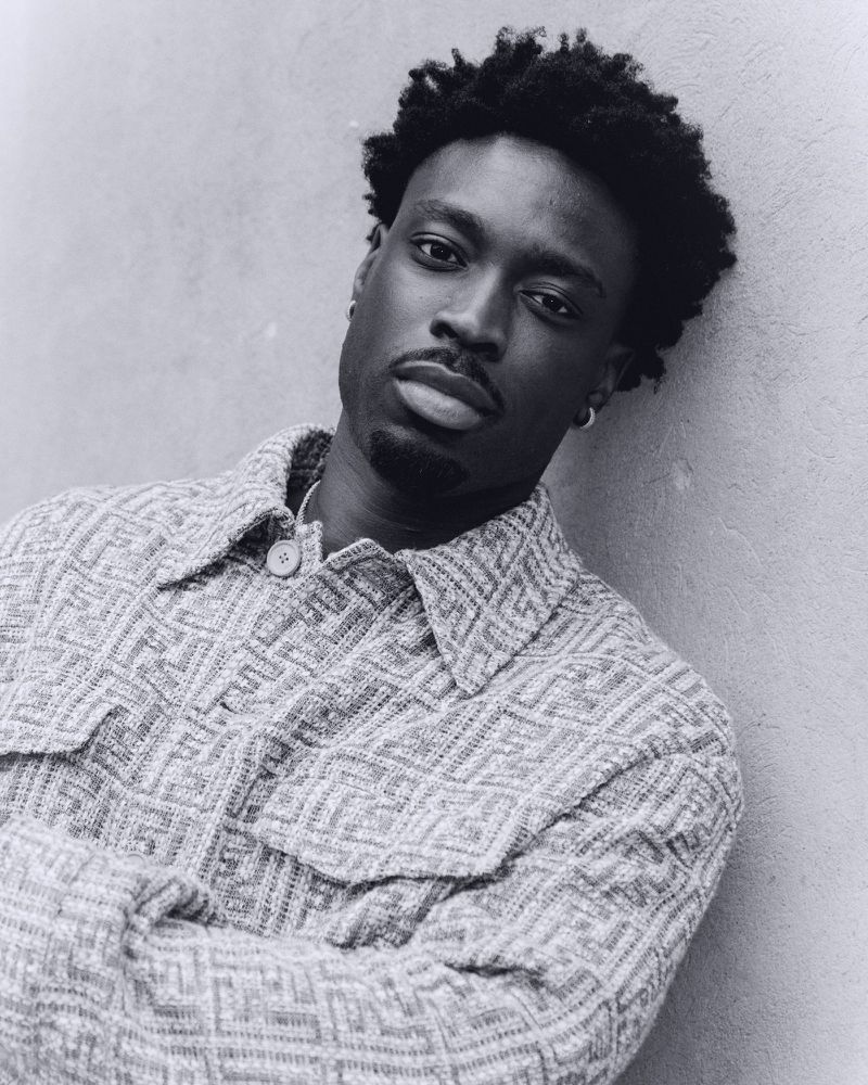 Portrait of a Black man with short curly hair, wearing a patterned button-down shirt, leaning against a gray wall indoors.